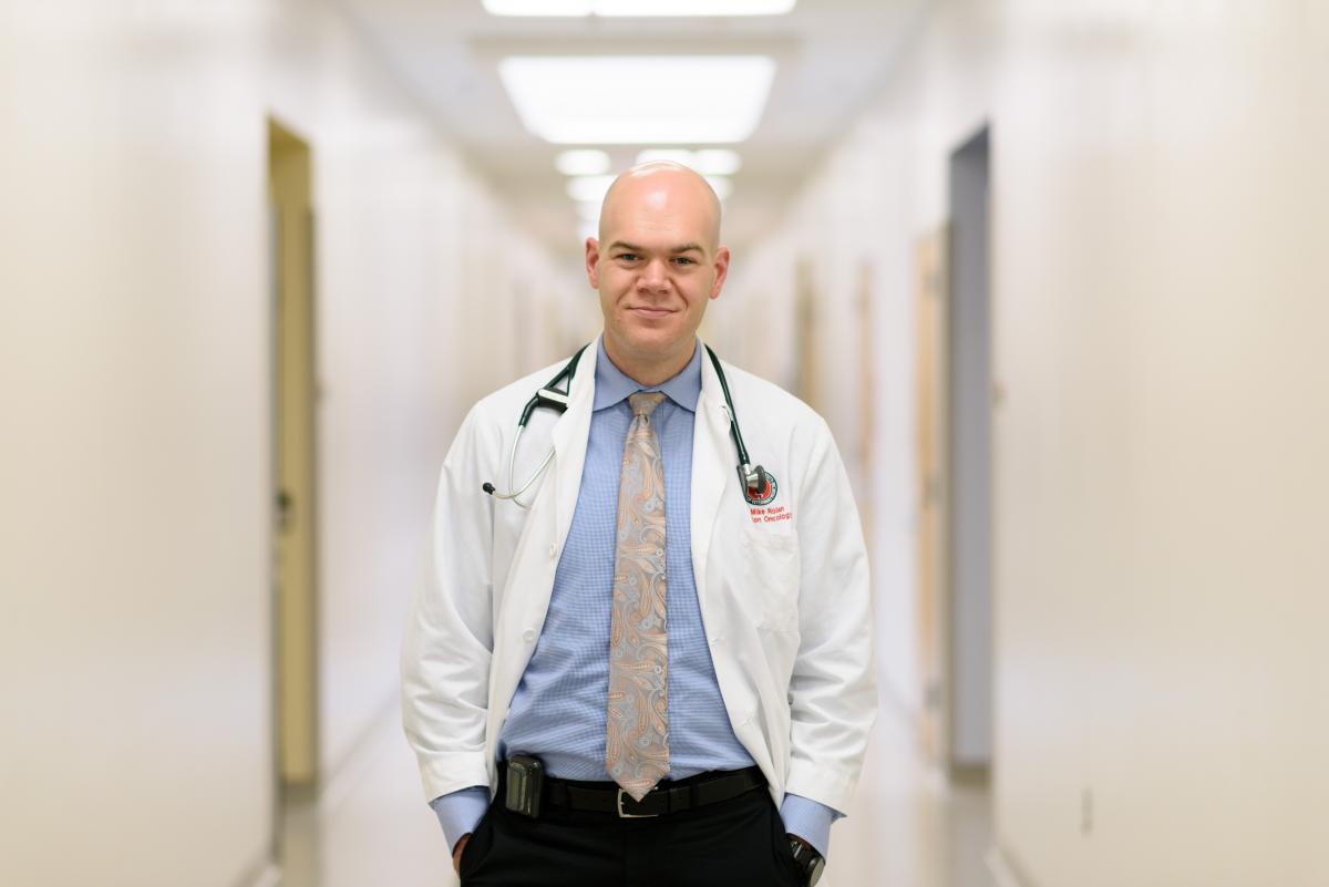 Images shows a bald light skinned man standing in a bright hallway wearing a white lab coat over a blue dress shirt and a tie with a stethescope around his neck and black pants.