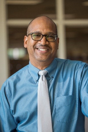 images shows smiling male with glasses and very short not shaved head, medium skin tone in a medium blue shirt with light blue tie