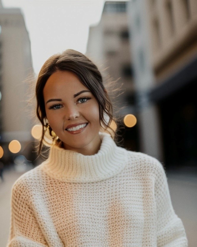 image shows the head and shoulders of a smiling light skinned woman with dark hair pulled back wearing a white mock turtleneck sweater