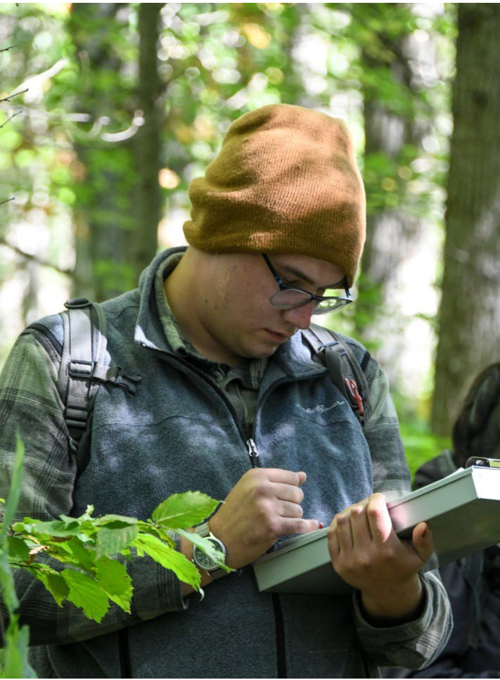 image shows the head and upper torso of a man wearing a brown winter slouch hat, blue fleece vest over a green plaid shirt and glasses looking down at a clipboard and making notes standing in a forest
