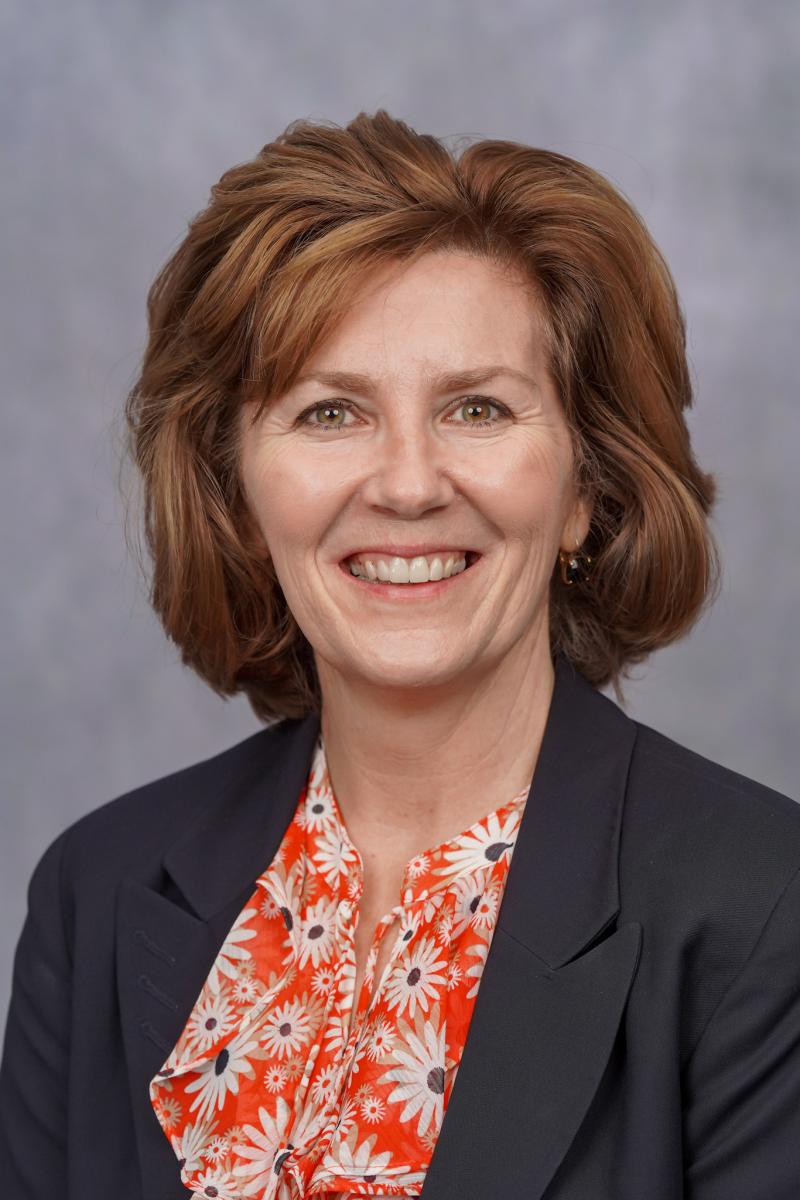 image shows the head and shoulders of a smiling light-skinned woman with medium length red-brown hair wearing a red/orange flowered blouse with a navy suit jacket