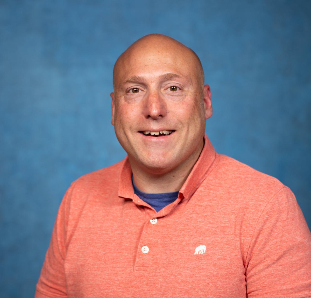 image shows the head and upper torso of a smiling light skinned bald man wearing a coral colored polo shirt