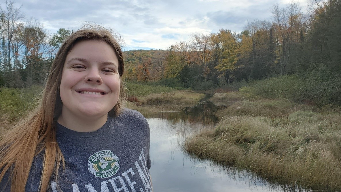 images shows a woman with long medium brown hair smiling, light skin and where a grey shirt with unreadable words on it in front of a river with grassy bank and trees in the far background