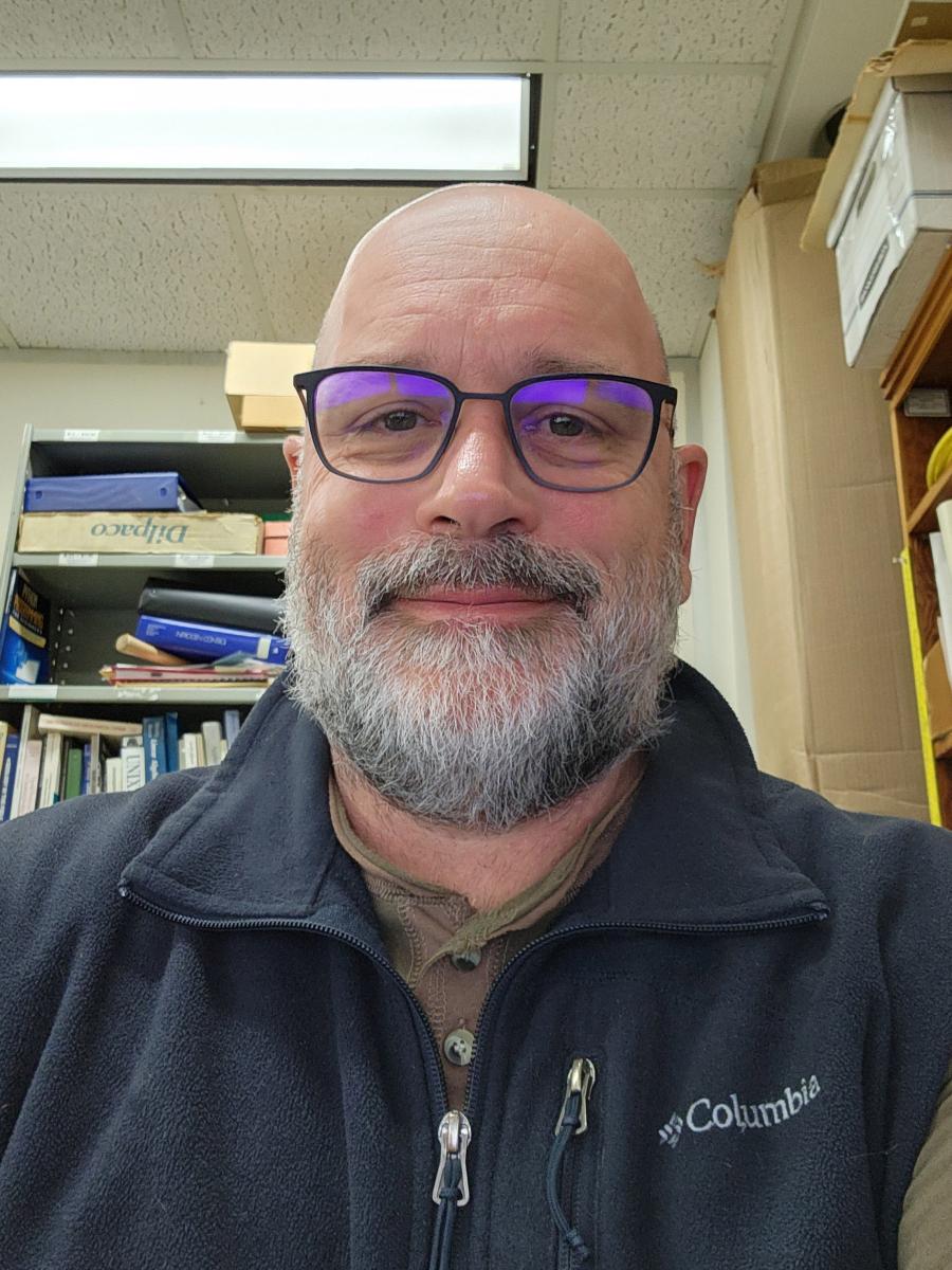 image shows the head and shoulders of a man with a bald head, gray beard and glasses wearing a navy blue fleece zipped up and a bemused smile